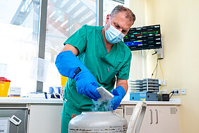 Eine Person in medizinischer Arbeitskleidung und mit Handschuhen und Mundschutz bekleidet entnimmt ein Behältnis aus einem großen Behälter, aus dem Dampf strömt.