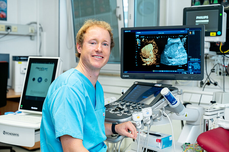 Eine Person in medizinischer Arbeitskleidung sitzt in einem Untersuchungsraum eines Krankenhauses. Rechts schräg hinter der Person befindet sich ein Monitor, der zwei Ultraschallbilder zeigt. Im Raum befinden sich weitere medizinische Geräte für Ultraschalluntersuchungen. Eine Person in medizinischer Arbeitskleidung sitzt in einem Untersuchungsraum eines Krankenhauses. Rechts schräg hinter der Person befindet sich ein Monitor, der zwei Ultraschallbilder zeigt. Im Raum befinden sich weitere medizinische Geräte für Ultraschalluntersuchungen.