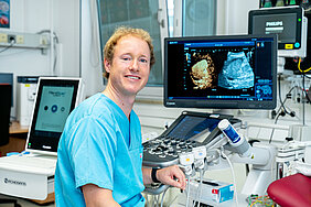 Eine Person in medizinischer Arbeitskleidung sitzt in einem Untersuchungsraum eines Krankenhauses. Rechts schräg hinter der Person befindet sich ein Monitor, der zwei Ultraschallbilder zeigt. Im Raum befinden sich weitere medizinische Geräte für Ultraschalluntersuchungen.
