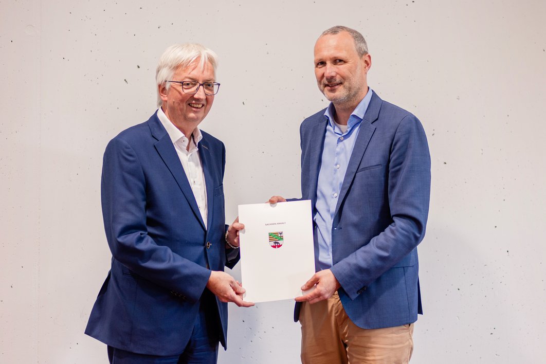 Zwei Männer mit blauem Hemd halten gemeinsam einen Umschlag in die Kamera und lächeln. Der linke Mann hat weiße Haare und trägt eine Brille. Der rechte Mann ist etwas größer, hat kurze graue Haare und trägt einen kurzgeschnittenen Vollbart. 