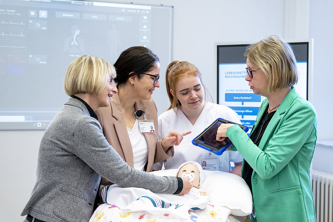 Vier Personen stehen um eine digitale Baby-Pflegepuppe herum. Eine Person erklärt die Funktionen auf einem Tablet.