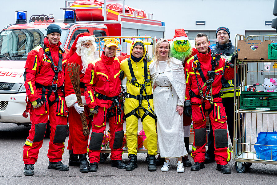 Eine Gruppe von Feuerwehrkräften in roten Einsatzanzügen steht lächelnd vor einem Feuerwehrfahrzeug. Neben ihnen sind Personen in Weihnachtskostümen zu sehen, darunter ein Weihnachtsmann, ein Engel, sowie zwei Personen im Pikachu- und Grinch-Kostüm. Rechts im Bild stehen Rollwagen mit Geschenken. Eine Gruppe von Feuerwehrkräften in roten Einsatzanzügen steht lächelnd vor einem Feuerwehrfahrzeug. Neben ihnen sind Personen in Weihnachtskostümen zu sehen, darunter ein Weihnachtsmann, ein Engel, sowie zwei Personen im Pikachu- und Grinch-Kostüm. Rechts im Bild stehen Rollwagen mit Geschenken.