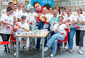 Eine Gruppe von insgesamt 15 Personen steht bzw. sitzt um einen Tisch, der voller Zahnpflegeutensilien ist, herum. Sie halten verschiedene Gegenstände in den Händen, darunter übergroße Zahnbürsten, Gebissmodelle, Übersichtskarten und Puppen.