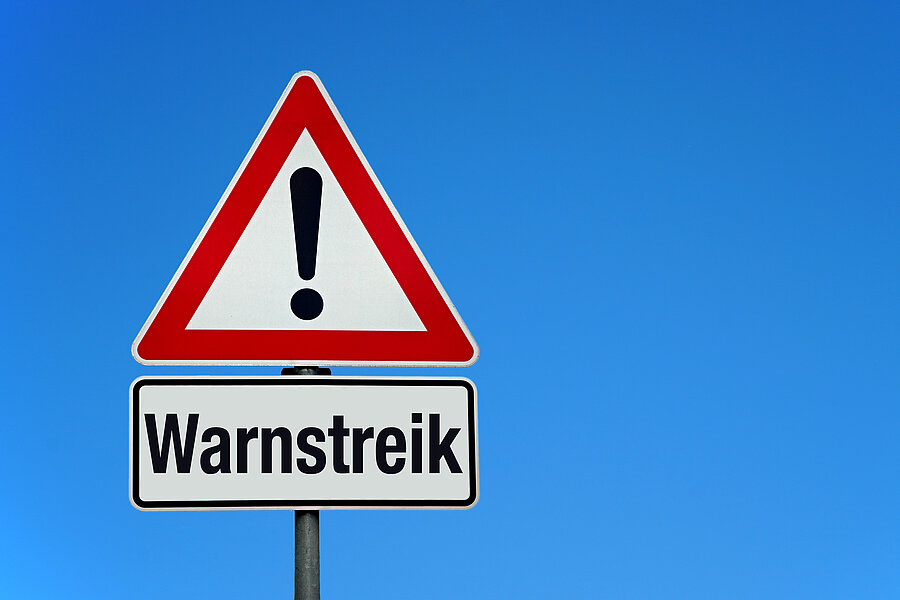 Vor blauem Himmel ein rotumrandetes Warnschild mit einem Ausrufezeichen. Darunter ein Schild, auf dem Warnstreik steht. Vor blauem Himmel ein rotumrandetes Warnschild mit einem Ausrufezeichen. Darunter ein Schild, auf dem Warnstreik steht.