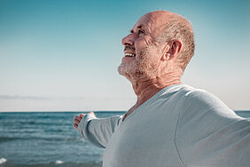 Ein Mann steht am Meer, breitet die Arme aus und lacht in die Landschaft. Er wirkt gelöst.