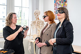 Zwei Personen tragen Brillen, mit denen sich Sehbeschwerden nachstellen lassen. Eine weitere Person ist mit ihnen im Gespräch und hält eine Brille in den Händen. Im Hintergrund steht ein Skelett, das für die Lehre genutzt wird.
