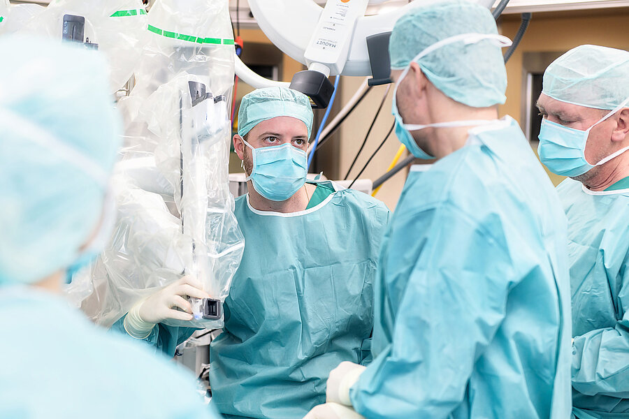 Vier Personen in OP-Kleidung stehen in einem Operationssaal in einer Gruppe zusammen. Links im Bild befindet sich einer der Arme des OP-Roboters da Vinci Xi. Vier Personen in OP-Kleidung stehen in einem Operationssaal in einer Gruppe zusammen. Links im Bild befindet sich einer der Arme des OP-Roboters da Vinci Xi.