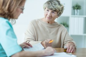 Eine ältere Frau sitzt frontal zur Kamera an einem Tisch. Sie blickt in das Gesicht einer anderen Frau mit Brille, mit der sie sich unterhält. Auf dem Tisch sind unscharf eine Medikamentenbox, ein Klemmbrett und Unterlagen zu erkennen. 