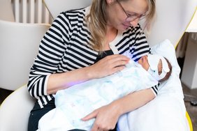 Eine Mutter hält ihr neugeborenes Baby im Arm, das in einem blau leuchtenden Einmal-Schlafsack mit kindlichem Aufdruck liegt. Das Baby trägt eine Binde über den Augen. Die Mutter schaut das Baby liebevoll an.