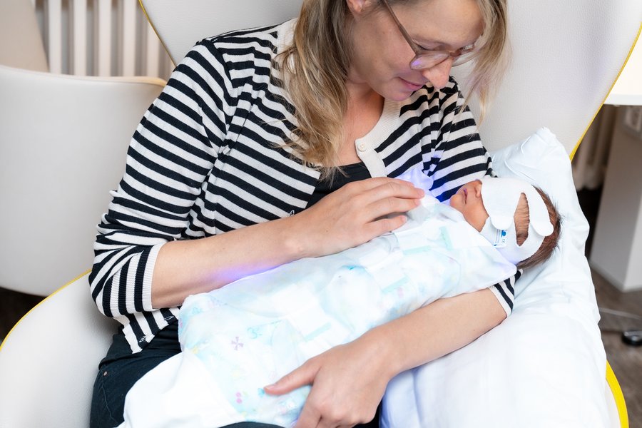 Eine Mutter hält ihr neugeborenes Baby im Arm, das in einem blau leuchtenden Einmal-Schlafsack mit kindlichem Aufdruck liegt. Das Baby trägt eine Binde über den Augen. Die Mutter schaut das Baby liebevoll an.  Eine Mutter hält ihr neugeborenes Baby im Arm, das in einem blau leuchtenden Einmal-Schlafsack mit kindlichem Aufdruck liegt. Das Baby trägt eine Binde über den Augen. Die Mutter schaut das Baby liebevoll an.