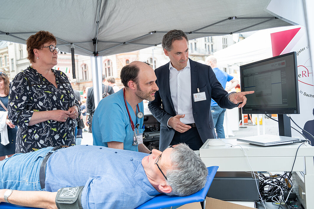 Bürgermeister Egbert Geier liegt auf einer Liege und trägt eine Blutdruckmanschette. Prof. Sedding zeigt Ministerin Petra Grimm-Benne und Dr. Mühlenweg etwas auf einem Monitor.