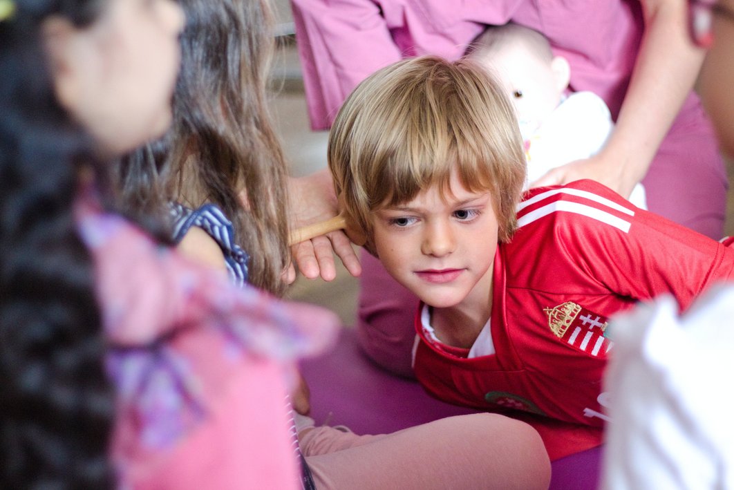 Nahaufnahme eines Jungen in rotem Fußballtrikot, der sein Ohr gegen ein hölzernes Hörrohr drückt, das von der Hand einer erwachsenen Person gestützt wird. Er schaut sehr konzentriert und aufmerksam. Um ihn herum sieht man in der Unschärfe weitere Personen.