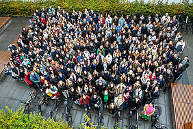 Das Gruppenbild aus der Vogelperspektive zeigt mehrere hundert Personen, die nach oben schauen. An dem nassen Boden und den dicken Jacken kann man erkennen, dass es zuvor geregnet hat. Im Hintergrund sind Büsche und Fahrräder zu sehen.