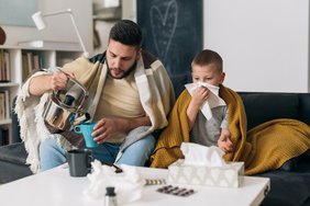 Ein Mann und ein Junge sitzen auf einem Sofa. Beide haben eine warme Decke um die Schultern. Der Mann gießt heißes Wasser aus einer Teekanne in eine Tasse. Der Junge putzt sich die Nase. Auf dem Tisch vor den beiden liegen verschiedene Tablettenschachteln und Taschentücher. Im Hintergrund sind ein Bücherregal, eine Lampe und ein Spiegel zu erkennen.