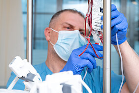 Eine Person in medizinischer Arbeitskleidung hält einen Blutbeutel für eine Stammzelltransplantation in den Händen. Die Person trägt eine Maske und Handschuhe.