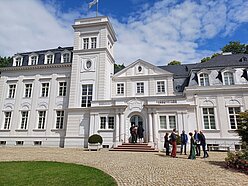 Außenansicht der großen weißen Villa, die den dritten Standort der HMU beherbergt, vor blauem Himmel mit weißen Wolken. Das Gebäude gliedert sich in mehrere Abschnitte, die unterschiedlich gestaltet sind und z.B. unterschiedlich hoch sind. Neben dem zentralen Haupteingang ragt ein Turm auf, die durchgängig weiße Fassade ist durch Zierelemente und Fenster gegliedert. das Dach ist anthrazitfarben. Im Vordergrund sind eine breite Kiesauffahrt sowie ein Stück Rasenfläche zu sehen. Die hallesche Delegation steht als Grüppchen rechts vor dem Haupteingang.