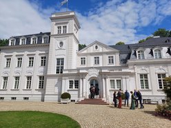 Außenansicht der großen weißen Villa, die den dritten Standort der HMU beherbergt, vor blauem Himmel mit weißen Wolken. Das Gebäude gliedert sich in mehrere Abschnitte, die unterschiedlich gestaltet sind und z.B. unterschiedlich hoch sind. Neben dem zentralen Haupteingang ragt ein Turm auf, die durchgängig weiße Fassade ist durch Zierelemente und Fenster gegliedert. das Dach ist anthrazitfarben. Im Vordergrund sind eine breite Kiesauffahrt sowie ein Stück Rasenfläche zu sehen. Die hallesche Delegation steht als Grüppchen rechts vor dem Haupteingang.