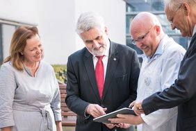 Dekanin Prof. Dr. Heike Kielstein, Wissenschaftsminister Prof. Dr. Armin Willingmann, Projektleiter Dr. Stefan Moritz und der Ärztliche Direktor Prof. Dr. Thomas Moesta (v.l.) stehen zusammen und schauen auf ein Tablet, das Dr. Moritz in der Hand hält. Prof. Willingmann und Prof. Moesta zeigen auf das Tablet.