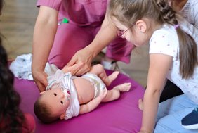 Ein Mädchen schaut aufmerksam auf eine Babypuppe, die auf einer Unterlage auf dem Boden liegt. Eine Person in beerenfarbener medizinischer Arbeitskleidung zieht der Puppe ein Jäckchen an.