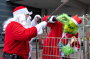 Ein als Grinch verkleideter Mensch steht in einem Gitterwagen, er hat Geschenke im Arm und duckt sich vor einem als Weihnachtsmann verkleideten Mann, der so tut, als ob er ihn mit der Rute haut.