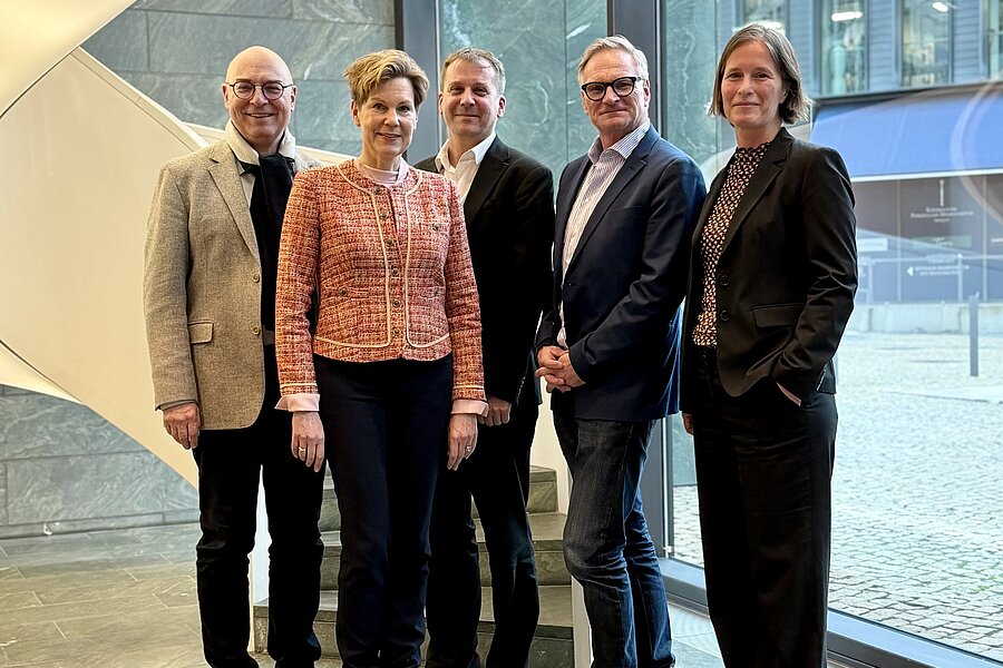 Fünf Personen stehen nebeneinander in einem Raum. Im Hintergrund ist eine beleuchtete, spiralförmige Treppe zu erkennen. Der Gebäudeteil auf der rechten Seite besteht vollständig aus Glas. Fünf Personen stehen nebeneinander in einem Raum. Im Hintergrund ist eine beleuchtete, spiralförmige Treppe zu erkennen. Der Gebäudeteil auf der rechten Seite besteht vollständig aus Glas.
