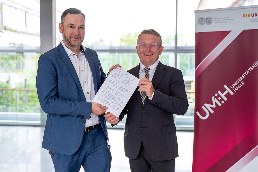 Zwei Männer in Änzügen stehen vor einem Roll-up mit dem Logo der UMH. Sie halten gemeinsam ein Schriftstück in die Kamera. Zwei Männer in Änzügen stehen vor einem Roll-up mit dem Logo der UMH. Sie halten gemeinsam ein Schriftstück in die Kamera.