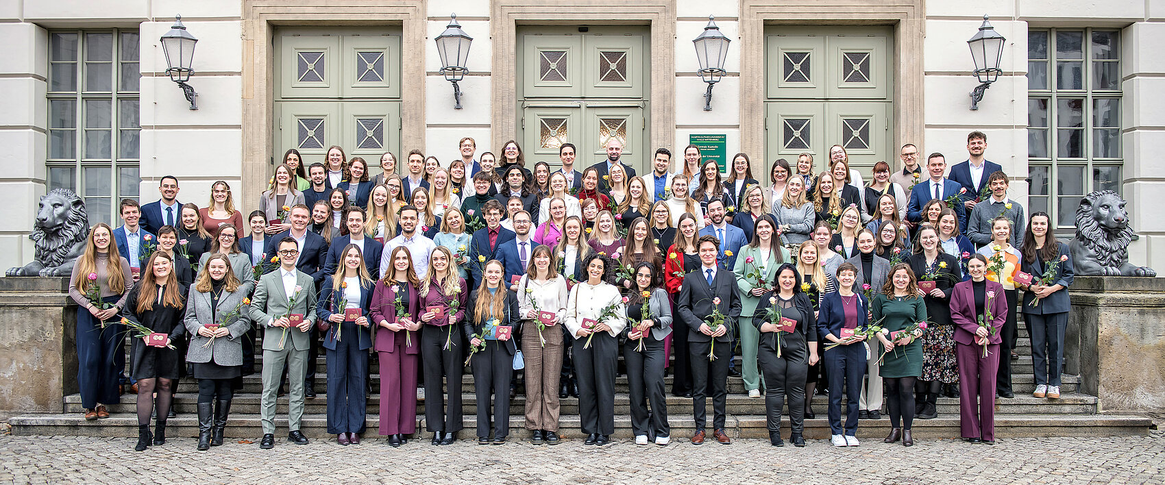 94 Personen stehen auf der Treppe vor dem Löwengebäude der Universität Halle-Wittenberg und lächeln in die Kamera. Sie halten jeweils ein kleines, dunkelrotes Couvert mit einem goldenen Siegel der Universität sowie eine orange-rote Rose in den Händen.
