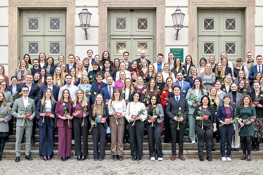 94 Personen stehen auf der Treppe vor dem Löwengebäude der Universität Halle-Wittenberg und lächeln in die Kamera. Sie halten jeweils ein kleines, dunkelrotes Couvert mit einem goldenen Siegel der Universität sowie eine orange-rote Rose in den Händen. 94 Personen stehen auf der Treppe vor dem Löwengebäude der Universität Halle-Wittenberg und lächeln in die Kamera. Sie halten jeweils ein kleines, dunkelrotes Couvert mit einem goldenen Siegel der Universität sowie eine orange-rote Rose in den Händen.