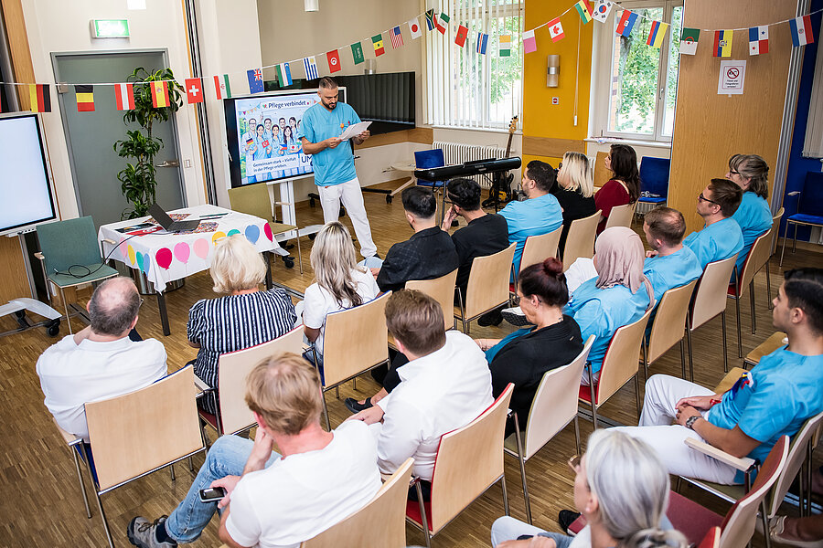 Teilnehmende eines Aktionstages sitzen in einem Raum. Vorne steht eine Person und erzählt etwas. Der Raum ist mit Girlanden aus Fahnen verschiedener Länder geschmückt. Teilnehmende eines Aktionstages sitzen in einem Raum. Vorne steht eine Person und erzählt etwas. Der Raum ist mit Girlanden aus Fahnen verschiedener Länder geschmückt.