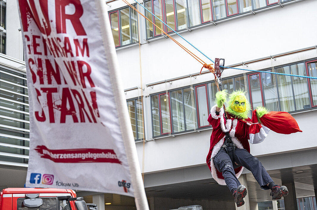 Ein als Grinch verkleideter Mitarbeiter der Feuerwehr seilt sich vom Universitätsklinikum Halle (Saale) ab. Links steht eine Fahne mit der Aufschrift "Nur gemeinsam sind wir stark".