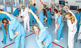 Eine Gruppe von Mitarbeitenden in einem Krankenhaus macht gemeinsam Sportübungen. Die Personen in blauer oder weißer medizinischer Arbeitskleidung stehen in Reihen und strecken einen Arm schräg nach oben, während die andere Hand an der Hüfte abgestützt ist. In dem Raum sieht man am Rand allerlei medizinische Geräte.