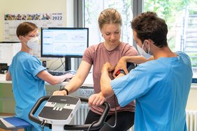 Drei Personen befinden sich in einem Trainingsraum. Eine junge Frau sitzt auf einem Heimtrainer und bekommt eine Blutdruckmanschette angelegt. Die dritte Person steht vor einem Monitor, auf dem unscharf mehrere Graphen zu erkennen sind, und blickt nach hinten zu den anderen beiden Personen.