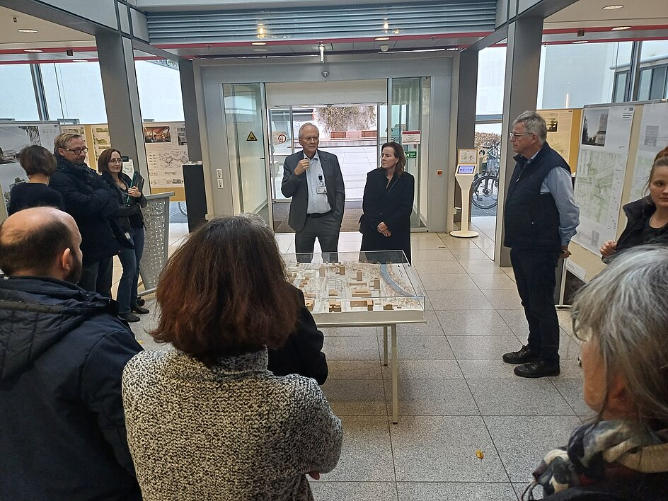 Im Rücken der Personen stehender Blick auf eine im Kreis angeordnete Gruppe von etwa 10 Menschen. In der Mitte des Kreises steht ein quadratischer Tisch, auf dem sich das maßstabsgetreue Standortmodell inklusive des Siegerentwurfs befindet. Auf der gegenüberliegenden Seite Blick auf 2 Menschen - den Ärztlichen Direktor Prof. Moesta und die Dekanin Prof. Kielstein - die gerade die Anwesenden begrüßen.