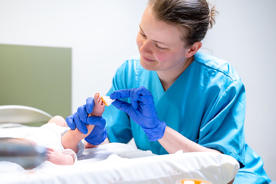 Eine Hebamme hält das Bein eines Babys in der Hand und bemalt den Fuß mit einem Tupfer und goldener Farbe. Eine Hebamme hält das Bein eines Babys in der Hand und bemalt den Fuß mit einem Tupfer und goldener Farbe.