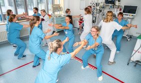 Eine Gruppe von Mitarbeitenden in einem Krankenhaus macht gemeinsam Sportübungen. Die Personen stehen sich gegenüber, haben die Knie gebeugt und die Arme ausgestreckt. In dem Raum sieht man am Rand allerlei medizinische Geräte.