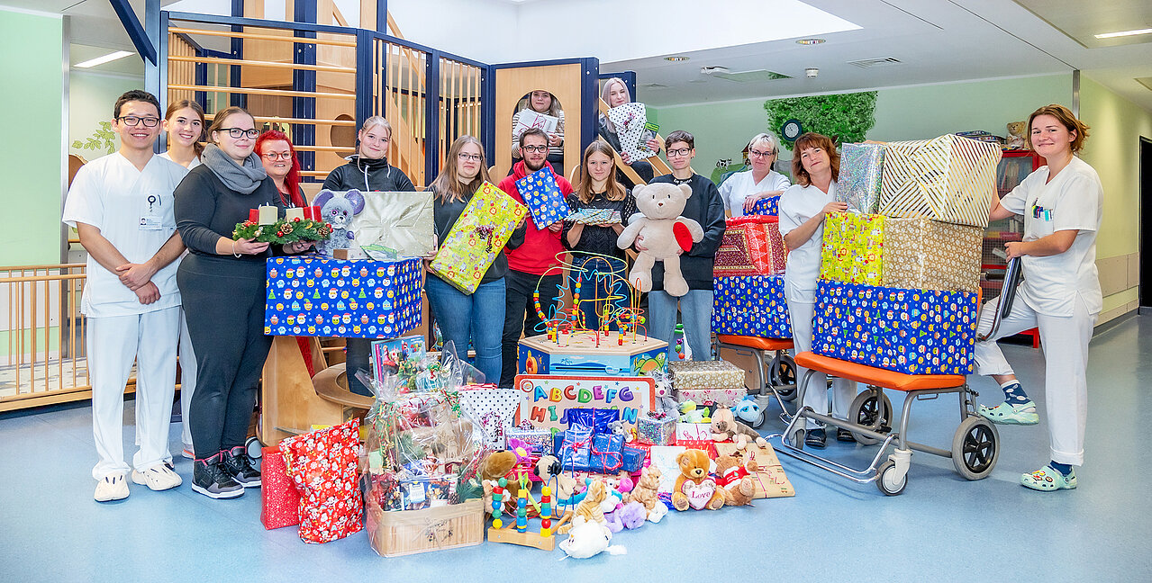 Eine große Gruppe von Menschen steht im Spielbereich einer Kinderstation in einem Krankenhaus. Vor der Gruppe stehen viele Geschenke. Einige Personen halten weitere Geschenke in den Händen. 