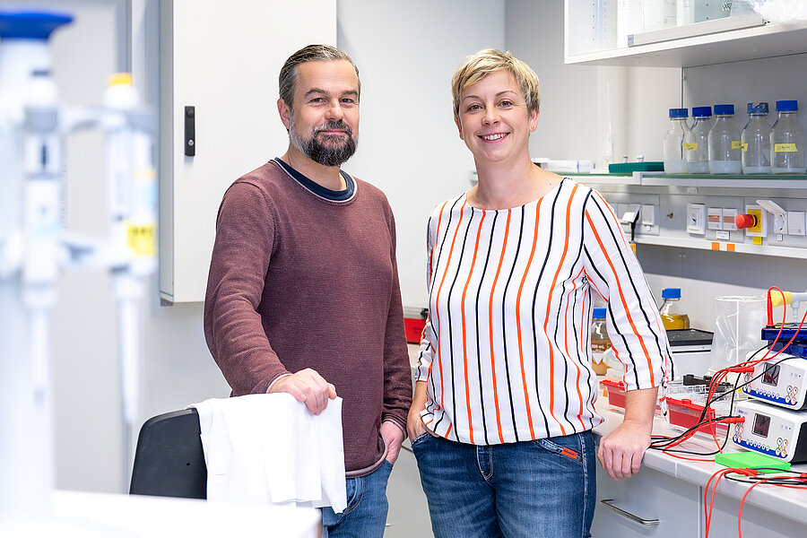 Zwei Personen stehen in einem weißen, hell erleuchteten Labor und lächeln in die Kamera. Links steht ein Mann in einem weinroten Pullover, der seine rechte Hand auf einem Stuhl abstützt. Auf der rechten Seite lehnt eine Frau mit gestreifter Bluse an einem Labortisch. Im Hintergrund und im Vordergrund sind verschiedene Laborgeräte wie Pipetten, Schottflaschen und Gelbäder zu sehen.  Zwei Personen stehen in einem weißen, hell erleuchteten Labor und lächeln in die Kamera. Links steht ein Mann in einem weinroten Pullover, der seine rechte Hand auf einem Stuhl abstützt. Auf der rechten Seite lehnt eine Frau mit gestreifter Bluse an einem Labortisch. Im Hintergrund und im Vordergrund sind verschiedene Laborgeräte wie Pipetten, Schottflaschen und Gelbäder zu sehen.