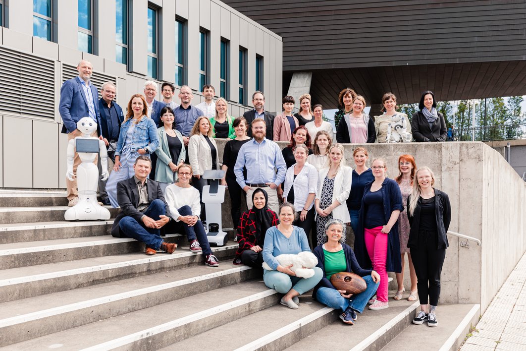 Mehr als 30 Personen stehen lächelnd und bei strahlender Sonne an einer Treppe vor dem Mitteldeutschen Multimediazentrum in Halle (Saale). Zwischen ihnen sind verschiedene digitale und assistive Technologien aufgebaut.