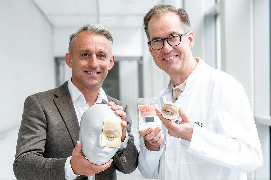 Zwei lächelnde Männer in einem hellen Klinikflur halten anatomische Augenmodelle in die Kamera: ein weißer Kopf mit aufgesetztem Lidmodell, ein transparenter Würfel mit Gewebeprobe und ein rundes Augenmodell mit Lidern. Zwei lächelnde Männer in einem hellen Klinikflur halten anatomische Augenmodelle in die Kamera: ein weißer Kopf mit aufgesetztem Lidmodell, ein transparenter Würfel mit Gewebeprobe und ein rundes Augenmodell mit Lidern.
