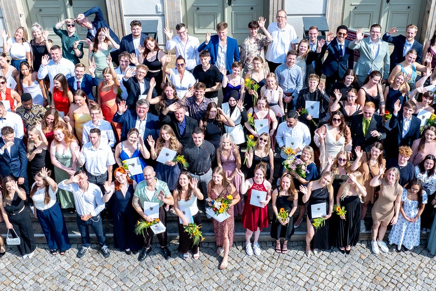 Drohnenaufnahme einer großen Gruppe von Menschen, die auf der Treppe vor dem Löwengebäude der Uni Halle steht. Viele sind festlich gekleidet und halten Zeugnisse und Blumen in den Händen. Die meisten schauen nach oben zur Drohne, viele winken in die Kamera.  Drohnenaufnahme einer großen Gruppe von Menschen, die auf der Treppe vor dem Löwengebäude der Uni Halle steht. Viele sind festlich gekleidet und halten Zeugnisse und Blumen in den Händen. Die meisten schauen nach oben zur Drohne, viele winken in die Kamera.