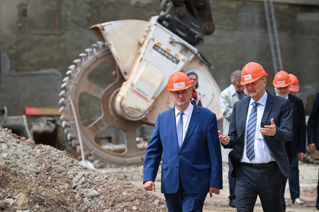 Ministerpräsident Reiner Haseloff (links) und Prof. Thomas Moesta gehen durch eine Baugrube. Beide tragen Anzüge und orangefarbene Helme. Dahinter befinden sich weitere Menschen. Im Hintergrund befinden sich Bauwerkzeuge.