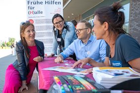 Halles Amtsärztin Christine Gröger und die Koordinatorin der Frühen Hilfen in Halle Julia Franke im Gespräch mit Prof. Dr. Roland Haase und Hebamme Katja Schumann. Sie sitzen an einem Tisch, auf dem ein rotes Banner mit UMH-Logo liegt. Im Hintergrund sieht man ein Roll-up von den Frühen Hilfen Halle.