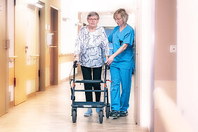 Eine ältere Dame und eine Person in medizinischer Arbeitskleidung gehen einen Krankenhausflur entlang. Die ältere Dame nutzt einen Rollator, die andere Person unterstützt sie.