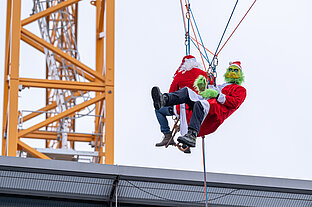 Zwei als Grinch und als Weihnachtsmann verkleidete Personen hängen an Sicherungsseilen in der Luft. Im Hintergrund ist ein Kran zu sehen.