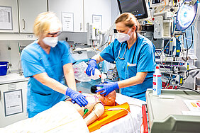 Zwei Personen in medizinischer Arbeitskleidung stehen in einem Behandlungsraum eines Krankenhauses. Beide tragen Masken und Handschuhe und trainieren eine Notfallsituation anhand einer Simulationspuppe. Im Hintergrund befinden sich Schränke und medizinische Geräte.