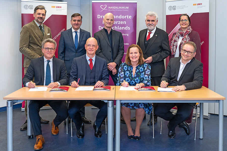 Gruppenfoto einer Vertragsunterzeichnung: Vier Personen sitzen an zwei Tischen und unterzeichnen Dokumente, hinter ihnen stehen weitere Beteiligte. Im Hintergrund sind Roll-ups des Universitätsklinikums Halle (UKH) und des Harzklinikums zu sehen Gruppenfoto einer Vertragsunterzeichnung: Vier Personen sitzen an zwei Tischen und unterzeichnen Dokumente, hinter ihnen stehen weitere Beteiligte. Im Hintergrund sind Roll-ups des Universitätsklinikums Halle (UKH) und des Harzklinikums zu sehen