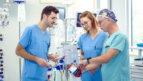 Zwei Männer und eine Frau (Mitte) in medizinischer Arbeitskleidung (grün und blau) stehen in einem Intensivstationszimmer. Sie haben Bauteile eines ECMO-Gerätes in den Händen und diskutieren miteinander..