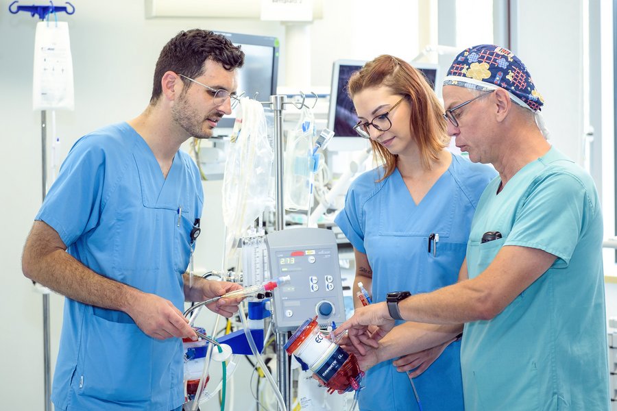 Zwei Männer und eine Frau (Mitte) in medizinischer Arbeitskleidung (grün und blau) stehen in einem Intensivstationszimmer. Sie haben Bauteile eines ECMO-Gerätes in den Händen und diskutieren miteinander..  Zwei Männer und eine Frau (Mitte) in medizinischer Arbeitskleidung (grün und blau) stehen in einem Intensivstationszimmer. Sie haben Bauteile eines ECMO-Gerätes in den Händen und diskutieren miteinander..