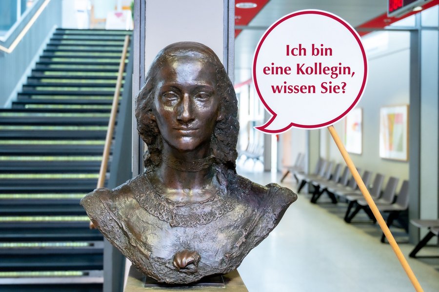 Auf einem Steinsockel steht die Büste einer Frau. Rechts neben der Büste wird eine Sprechblase aus Papier mit einem Stock in die Höhe gehalten. In der Sprechblase steht „Ich bin eine Kollegin, wissen Sie“.  Auf einem Steinsockel steht die Büste einer Frau. Rechts neben der Büste wird eine Sprechblase aus Papier mit einem Stock in die Höhe gehalten. In der Sprechblase steht „Ich bin eine Kollegin, wissen Sie“.