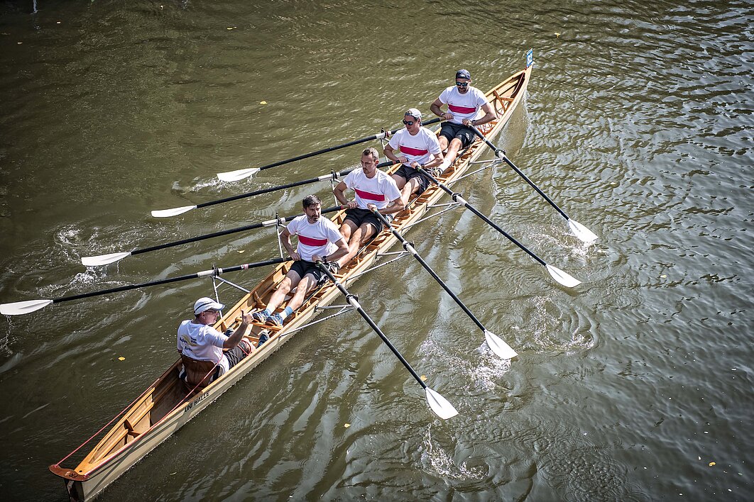 Vier Personen und ein Steuermann rudern in einem Ruderboot auf der Saale in Halle. Das Foto wurde von oben aufgenommen, das Boot rudert diagonal durchs Bild.